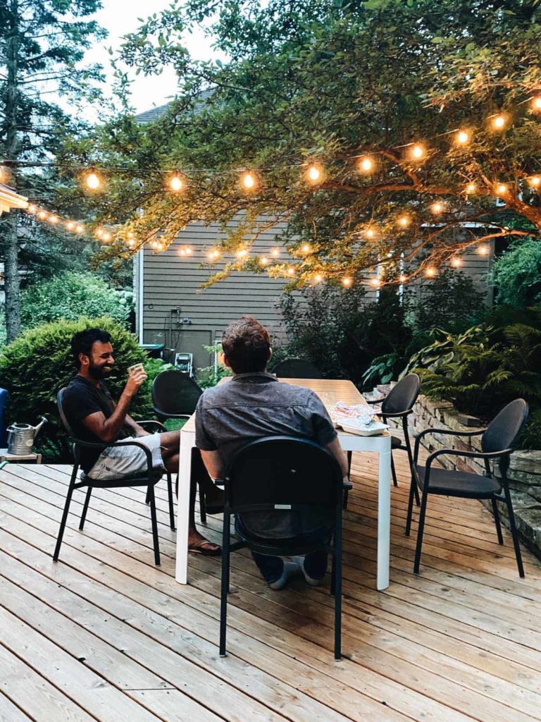 Two men sitting at a patio table laughing underneath overhead string lights