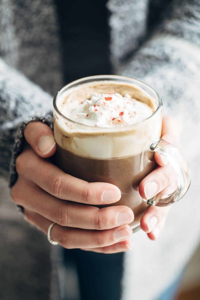 two hands holding a glass mug of hot chocolate with whipped cream and candy cane crumbles on top
