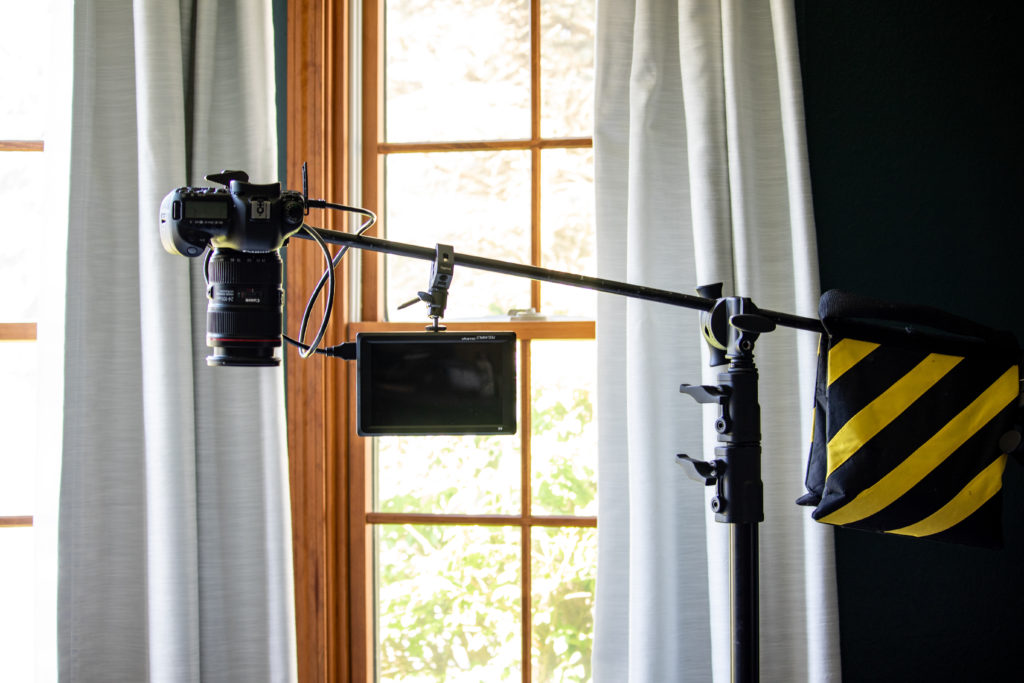An Impact C-Stand with Sliding Leg Kit with a sandbag on one end and a DSLR camera and FeelWorld Ultra-Bright Monitor on the other end