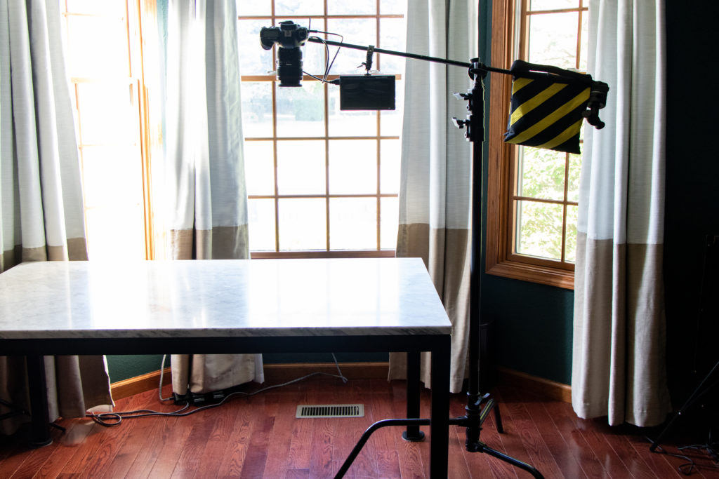 An Impact C-Stand with Sliding Leg Kit with a sandbag on one end and a DSLR camera and FeelWorld Ultra-Bright Monitor on the other end, set-up over a marble table for an overhead photograph.