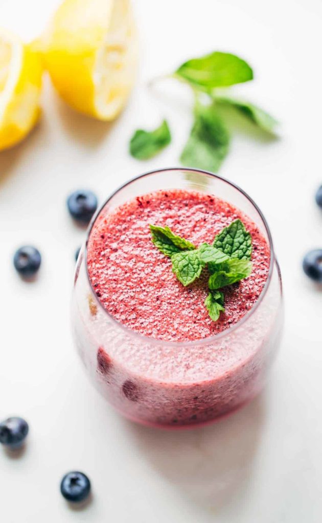 A glass filled with blueberry lemonade and topped with mint, surrounded by blueberries, mint, and lemons.