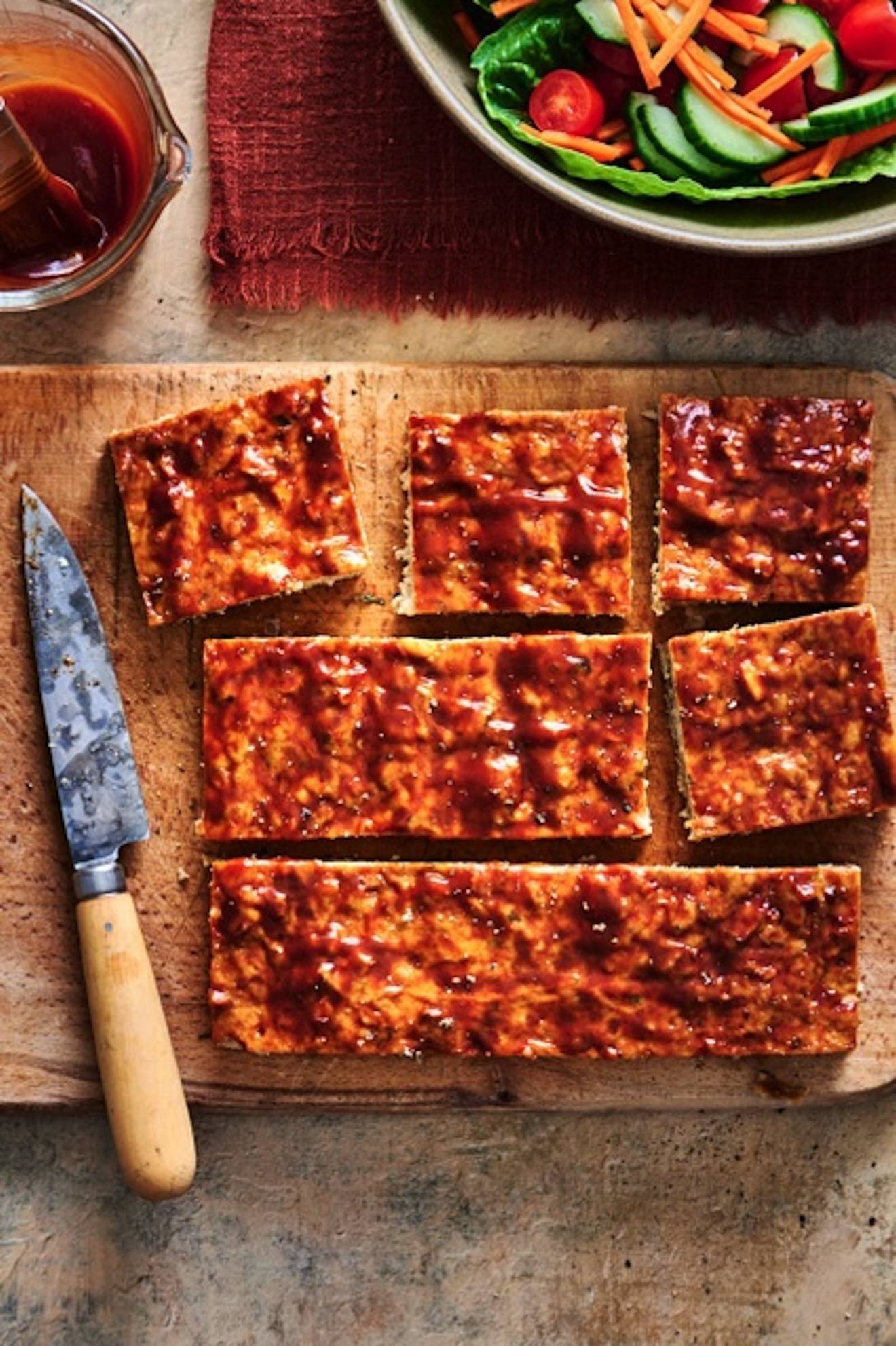 A photograph of meatloaf cut into squares on a cutting board with a green salad in the upper right corner.