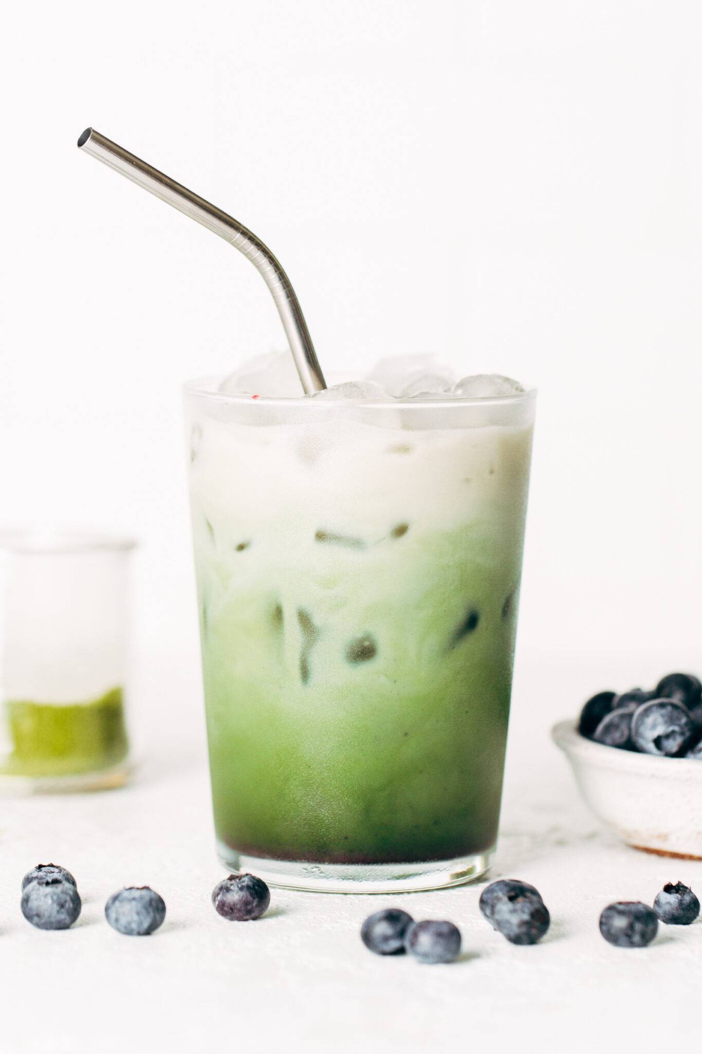 A photograph of a blueberry matcha latte in a glass with a metal straw.